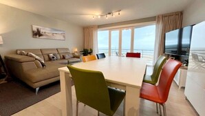 A modern living room with a dining area, a sofa, a TV, and a balcony with a view. - Apartment in Wenduine With Stunning Sea Views (De Haan)