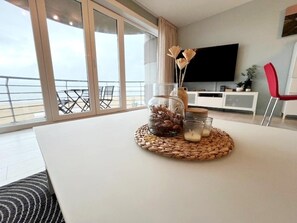 Een moderne woonkamer met een groot raam, een flatscreen televisie en een eethoek met een witte tafel en een geweven placemat.