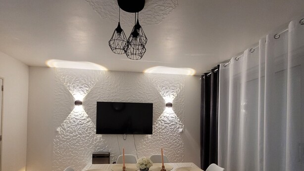 Un coin repas avec une table ronde, des chaises blanches, un téléviseur à écran plat fixé au mur et des luminaires suspendus.