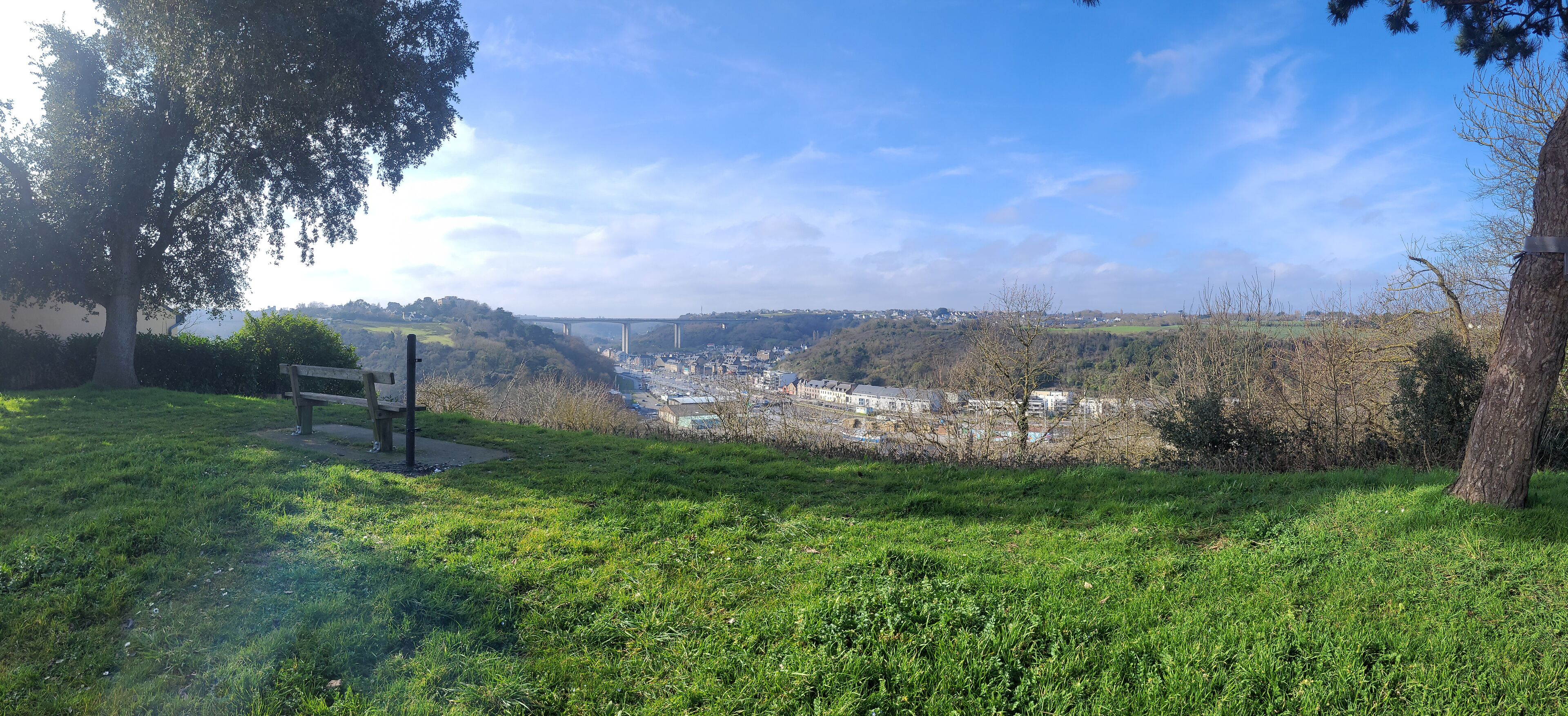 Zona verde con un banco, árboles y una vista distante de un puente sobre un valle.