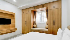 Un dormitorio con cama, armario de madera, televisor empotrado, ventana con cortinas y un banco.