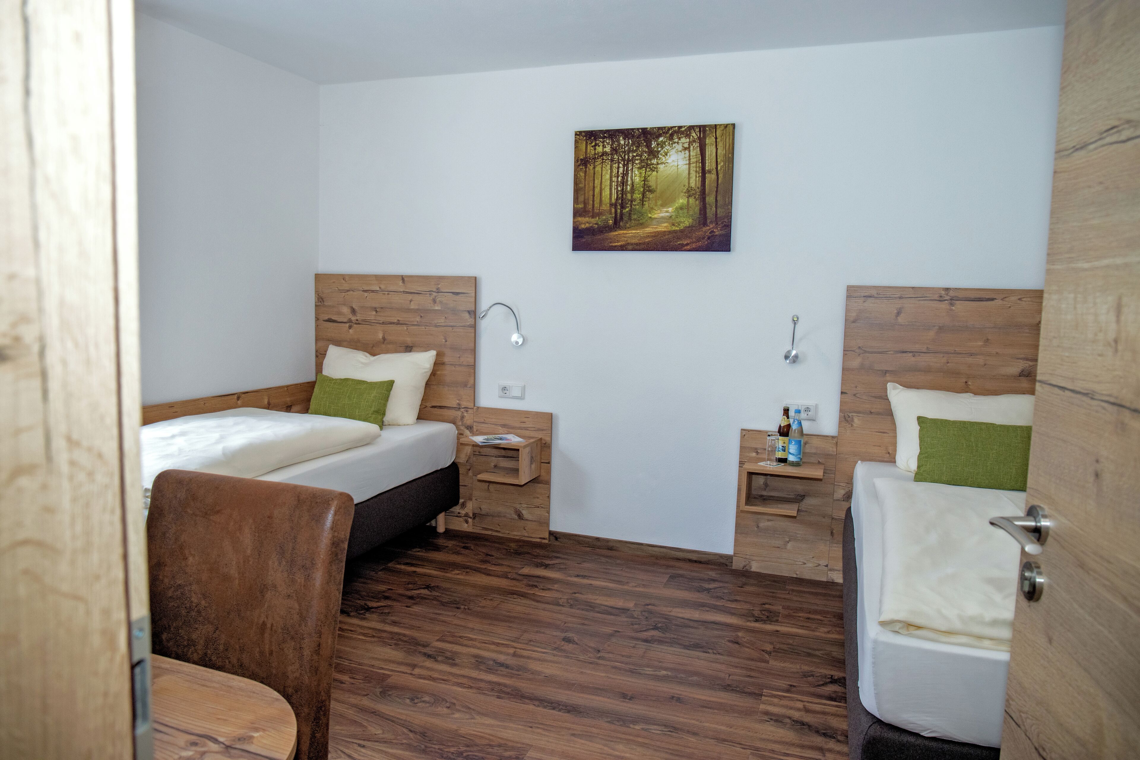 A bedroom with two beds, a wooden floor, a painting of a forest, and a small bedside table with a bottle.