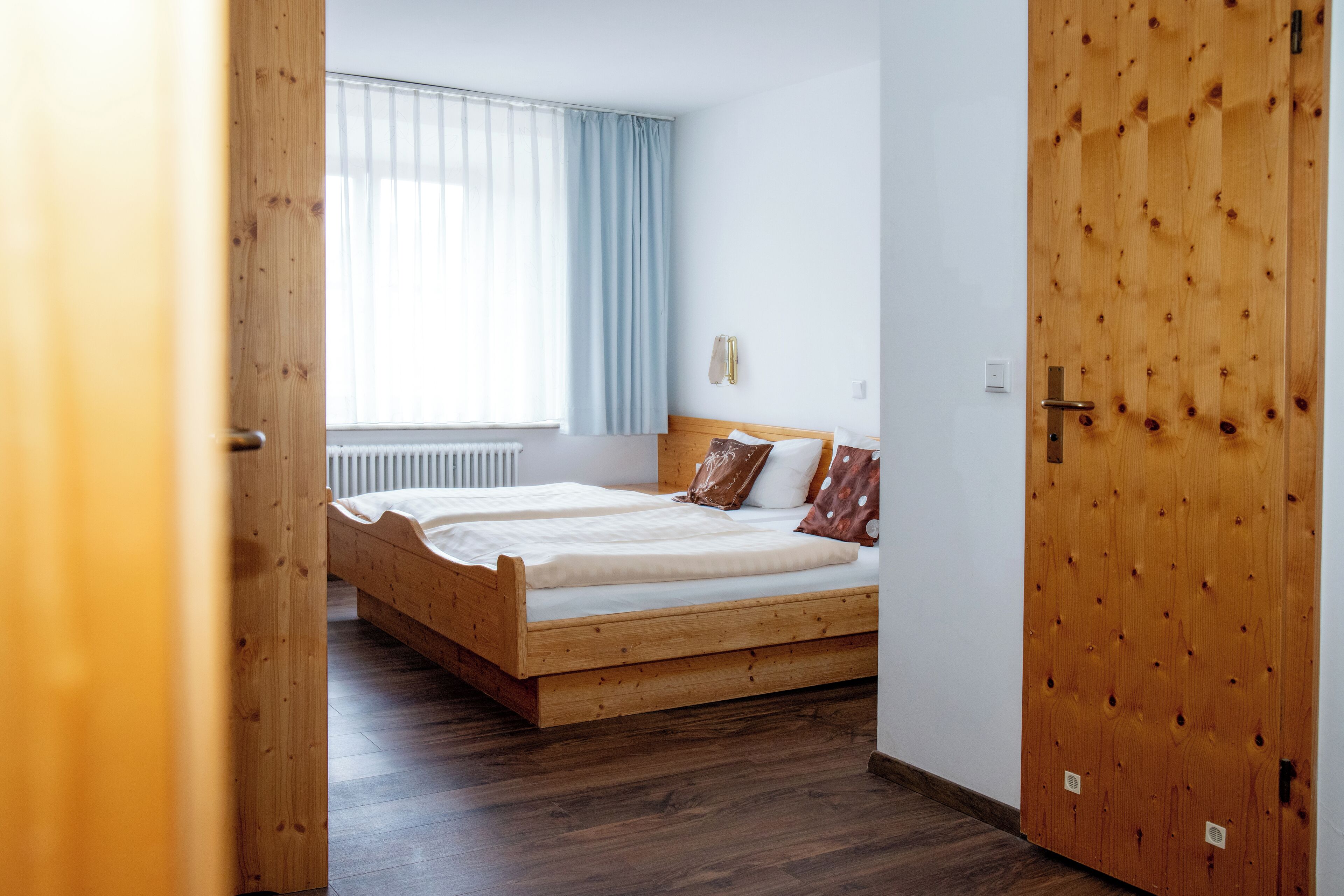 A bedroom with a wooden bed, a window with curtains, and a wooden door.