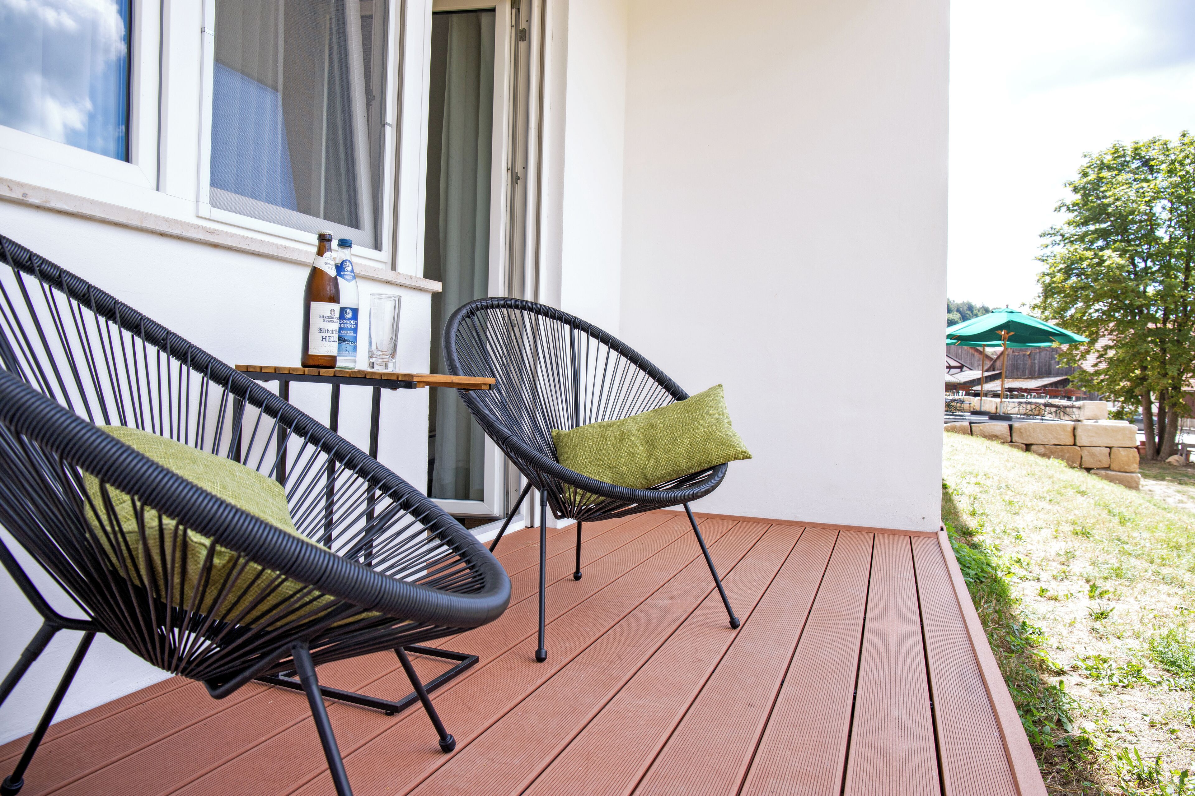 A wooden deck with two black wicker chairs, one with a green cushion, and a bottle of water and glass on a small table.