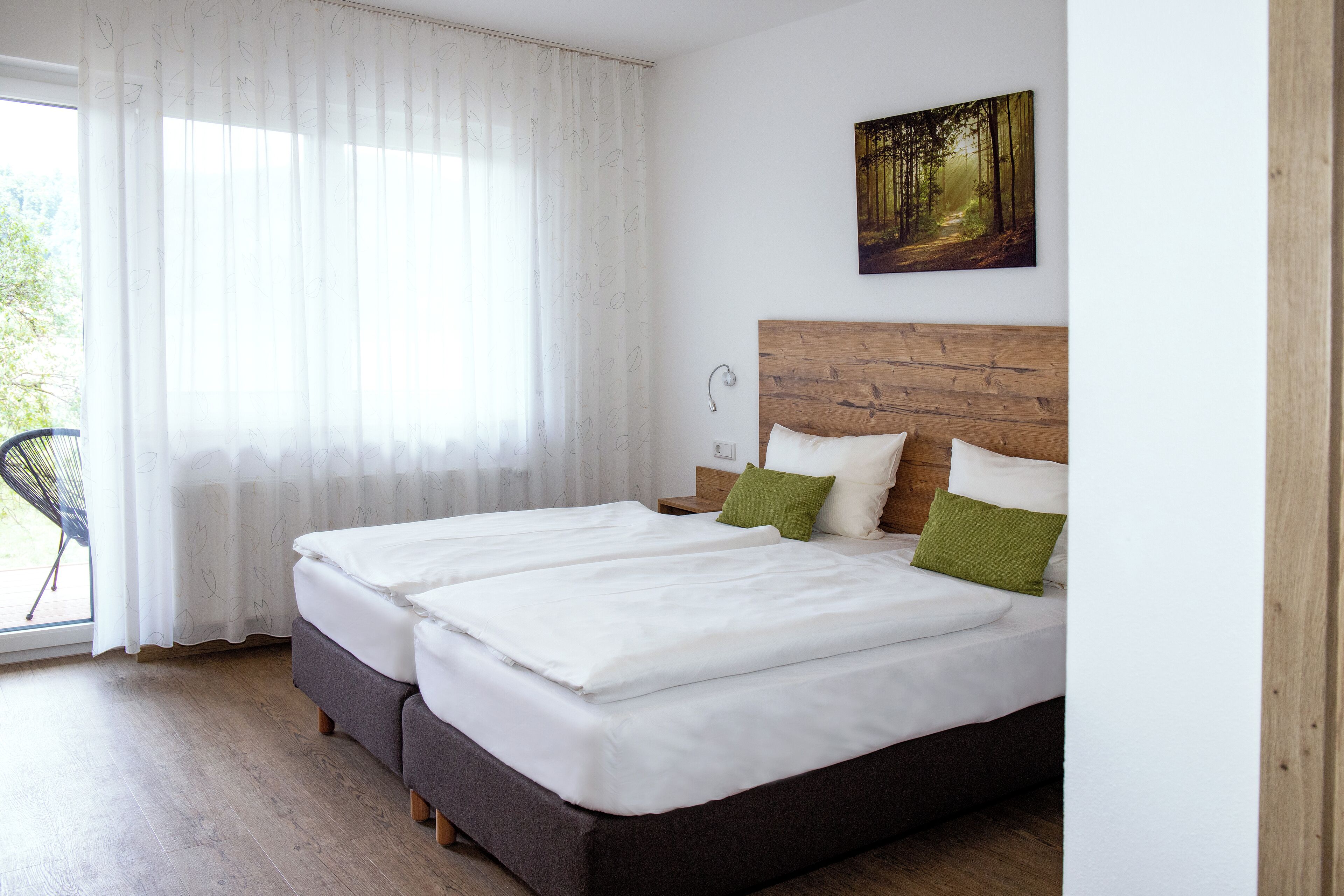 Un lit double avec une literie blanche et des oreillers verts, une tête de lit en bois et un tableau sur le thème de la forêt accroché au mur.