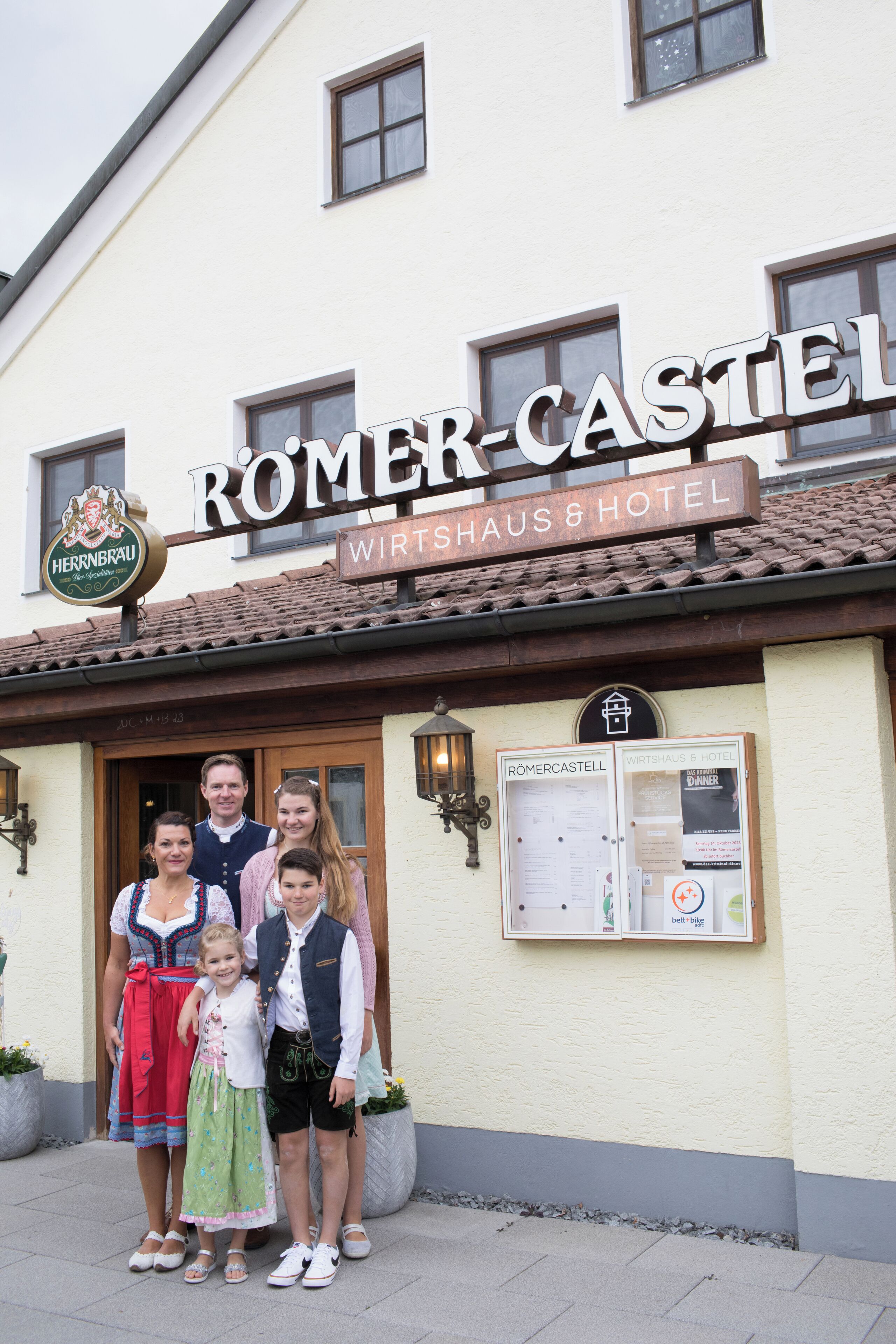 Uma família em frente a um prédio com uma placa que diz 'ROMER-CASTEL Wirtshaus & Hotel'.
