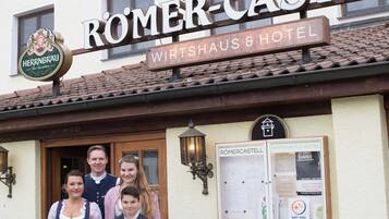 Uma família em frente a um prédio com uma placa que diz 'ROMER-CASTEL Wirtshaus & Hotel'.