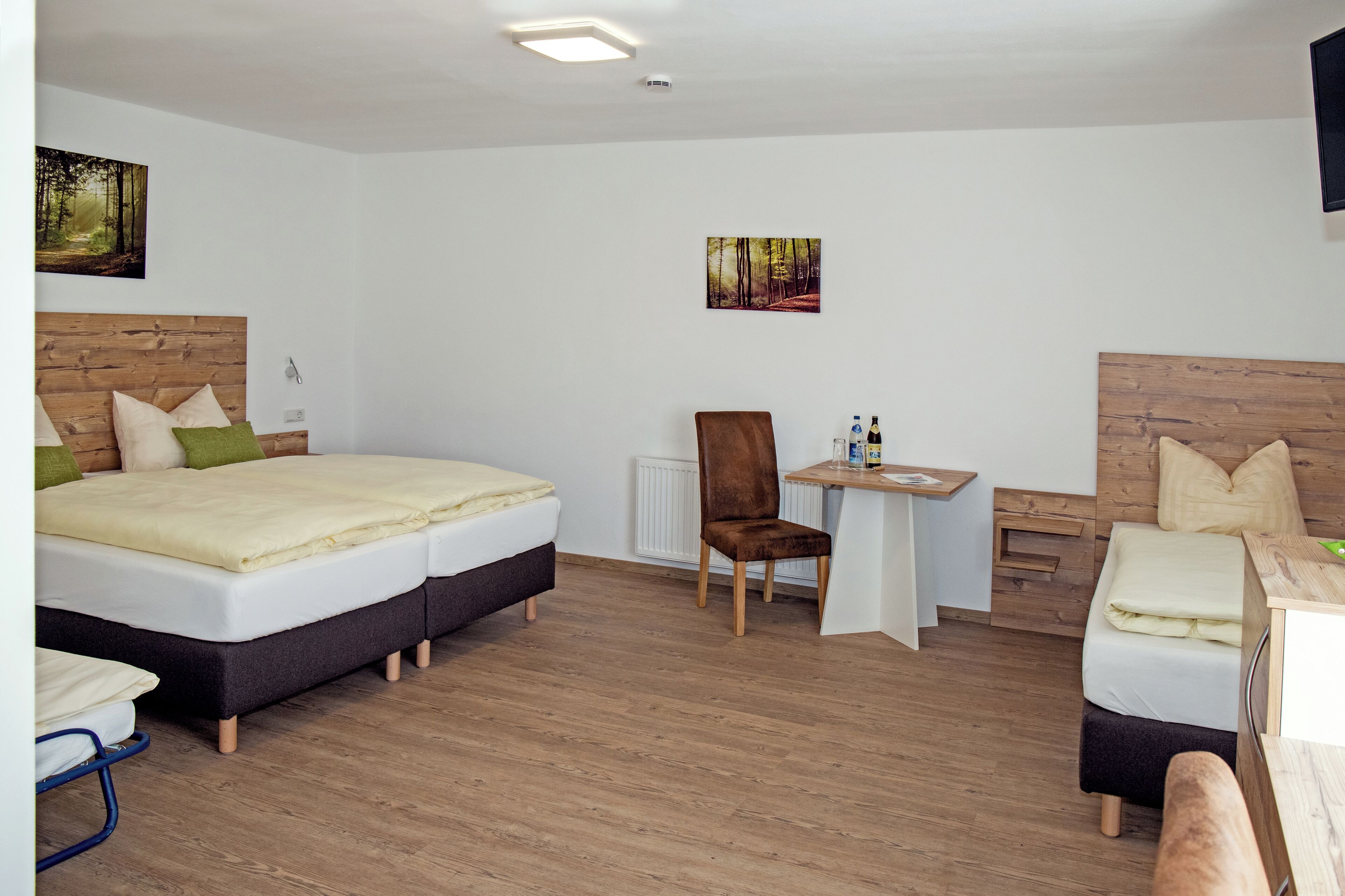 Une chambre d’hôtel moderne avec un grand lit, un bureau et une chaise. Deux tableaux ornent les murs, et une petite table porte une bouteille.