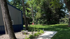 A paved pathway with gravel and small plants, surrounded by trees and a building with a blue and white exterior.