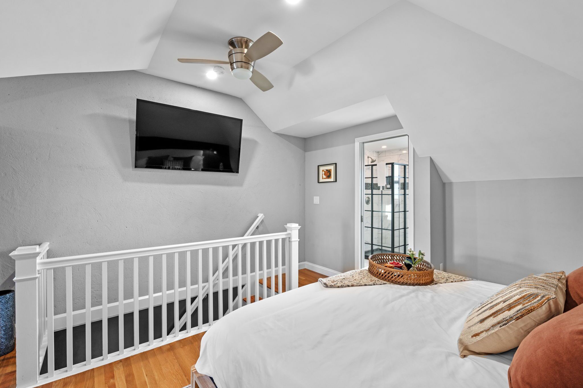 A bedroom with a white bed, a ceiling fan, a small black TV mounted on the wall, and a window with a view of the outside.