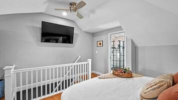 A bedroom with a white bed, a ceiling fan, a small black TV mounted on the wall, and a window with a view of the outside.