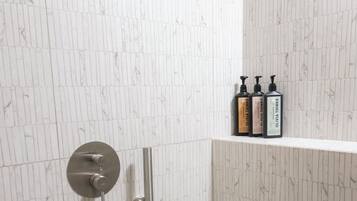 Ducha con manguera de mano y grifería empotrada. Hay un estante con artículos de aseo.