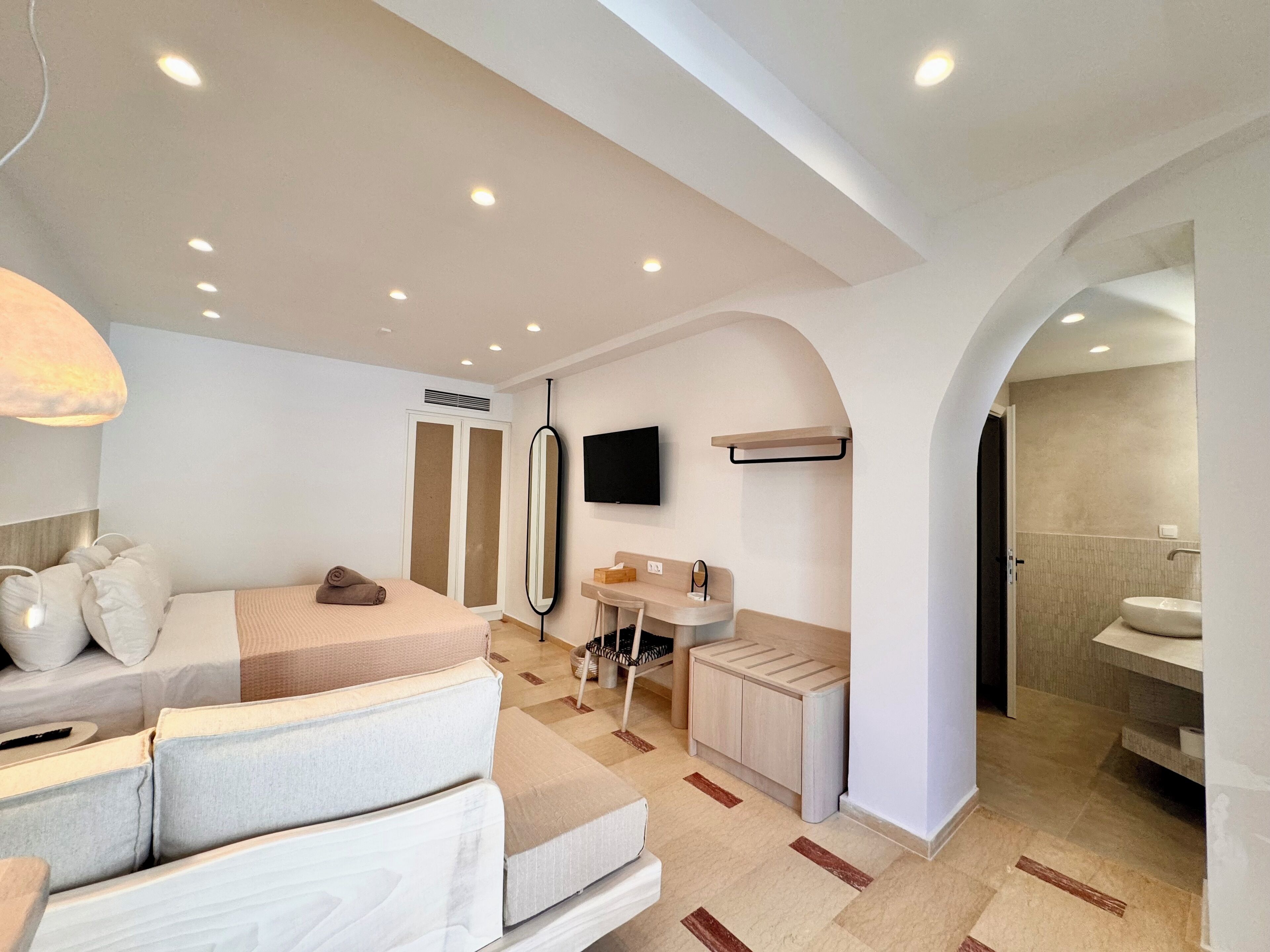 Un dormitorio moderno con cama, mesita de noche, televisor, armario y baño con lavabo y espejo.