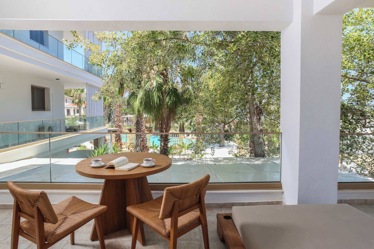 Una camera moderna con un balcone, un tavolo rotondo con sedie e vista su una piscina e vegetazione.