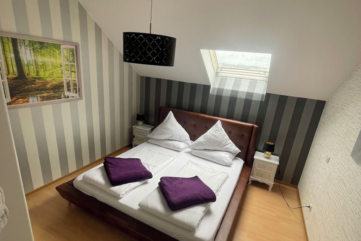 Ein Schlafzimmer mit einem Bett, einem Nachttisch, einer Lampe, einem Gemälde und einem Dachfenster.
