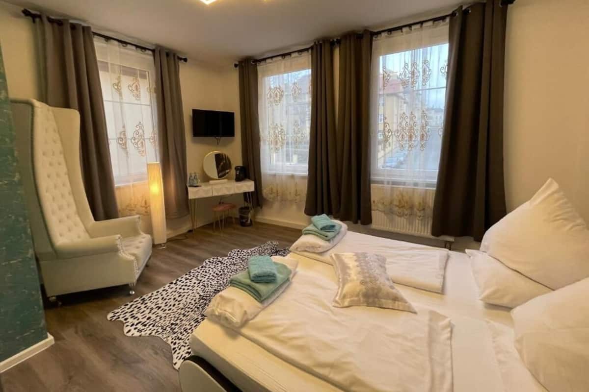 Ein Hotelzimmer mit einem großen Bett, einem Sessel, einem Fernseher und einem Fenster mit Vorhängen.