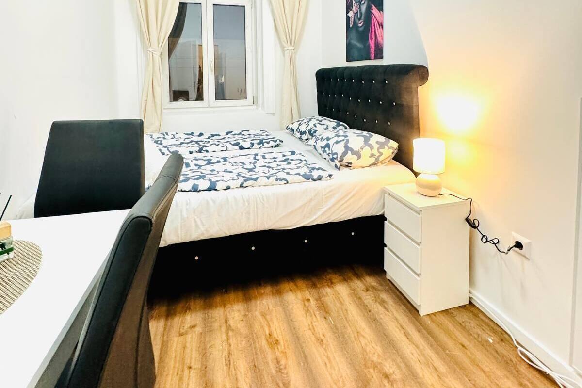 Ein Schlafzimmer mit Bett, Nachttisch mit Lampe, Stuhl und einem Fenster mit Vorhängen.