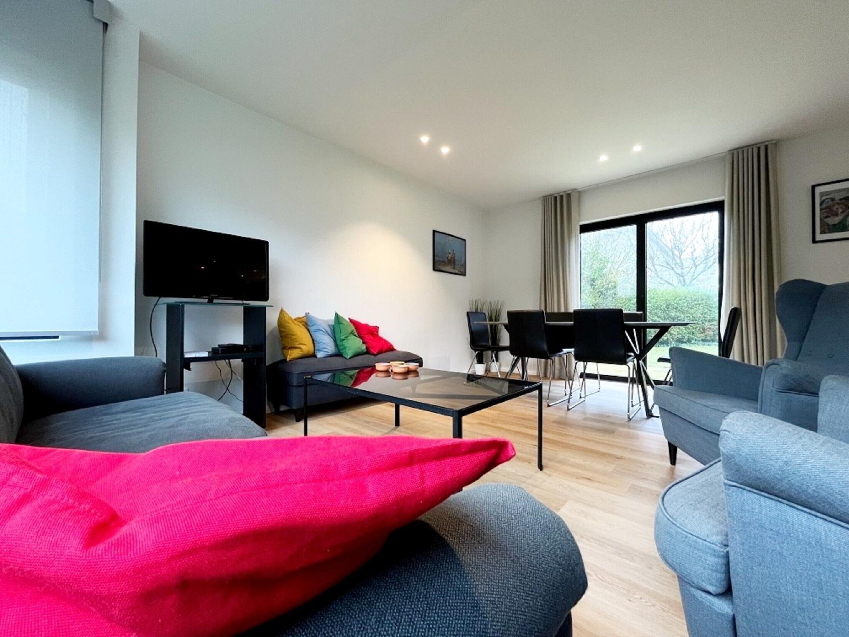 A modern living room with a grey sofa, a red throw blanket, a television, a dining table with chairs, and a large window with a view of greenery.