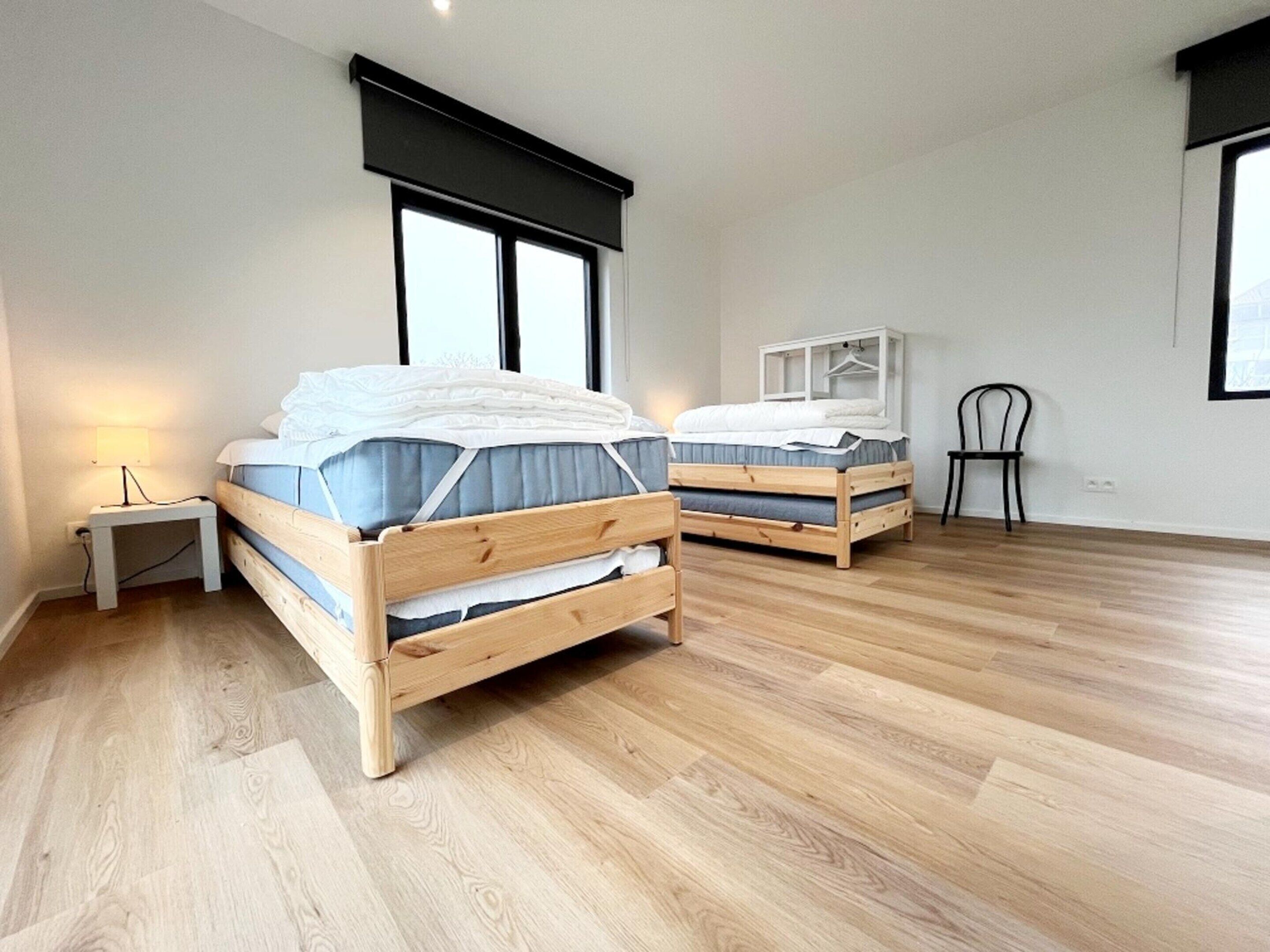 A modern bedroom with two wooden beds, a nightstand, a chair, and large windows.