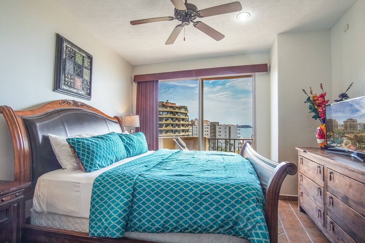 A bedroom with a bed, a ceiling fan, a nightstand, a dresser, and a view of buildings through a window.