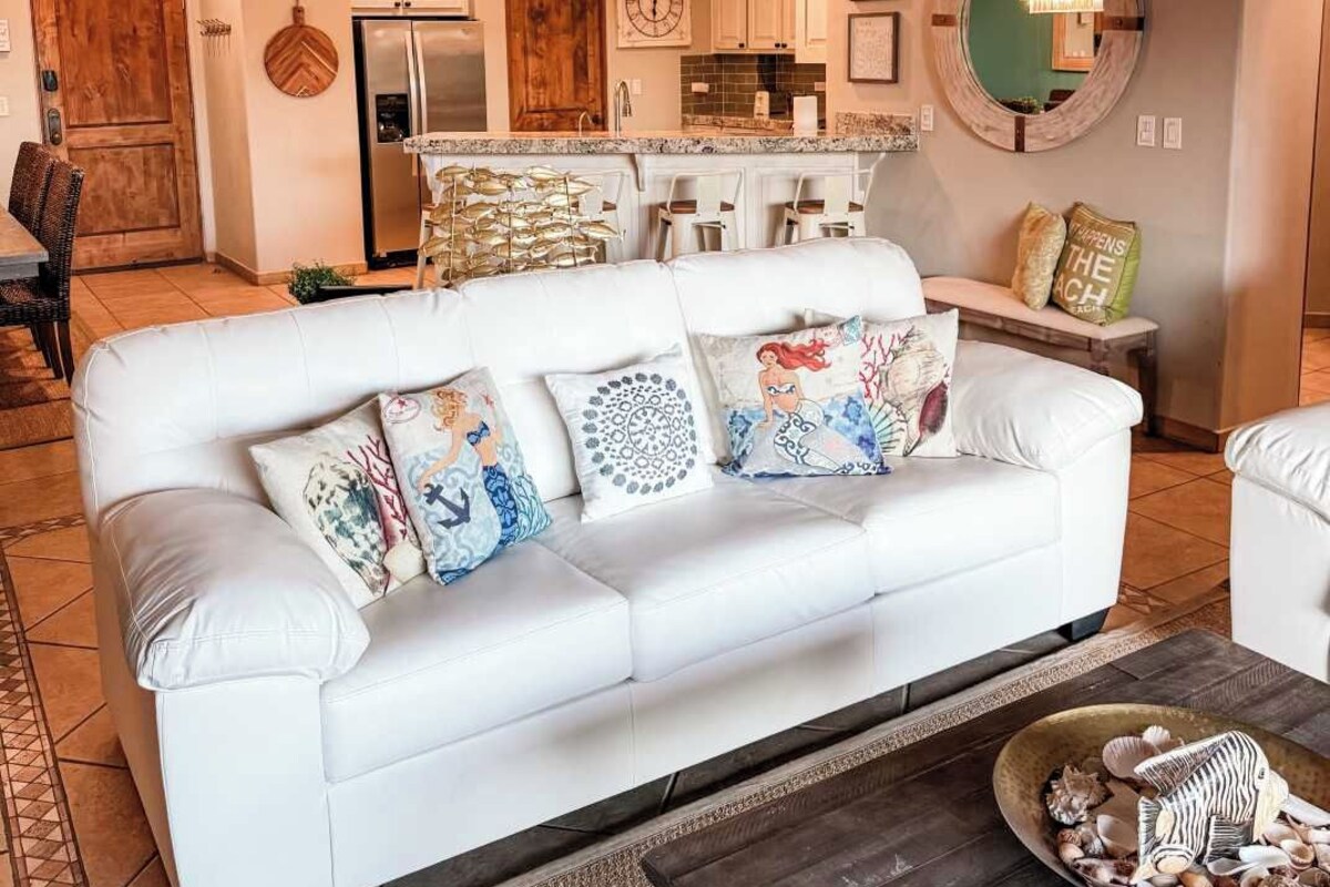 Een moderne woonkamer met een witte bank, decoratieve kussens, een bijzettafeltje met schelpen, een keuken op de achtergrond en een opvallend plafondontwerp.