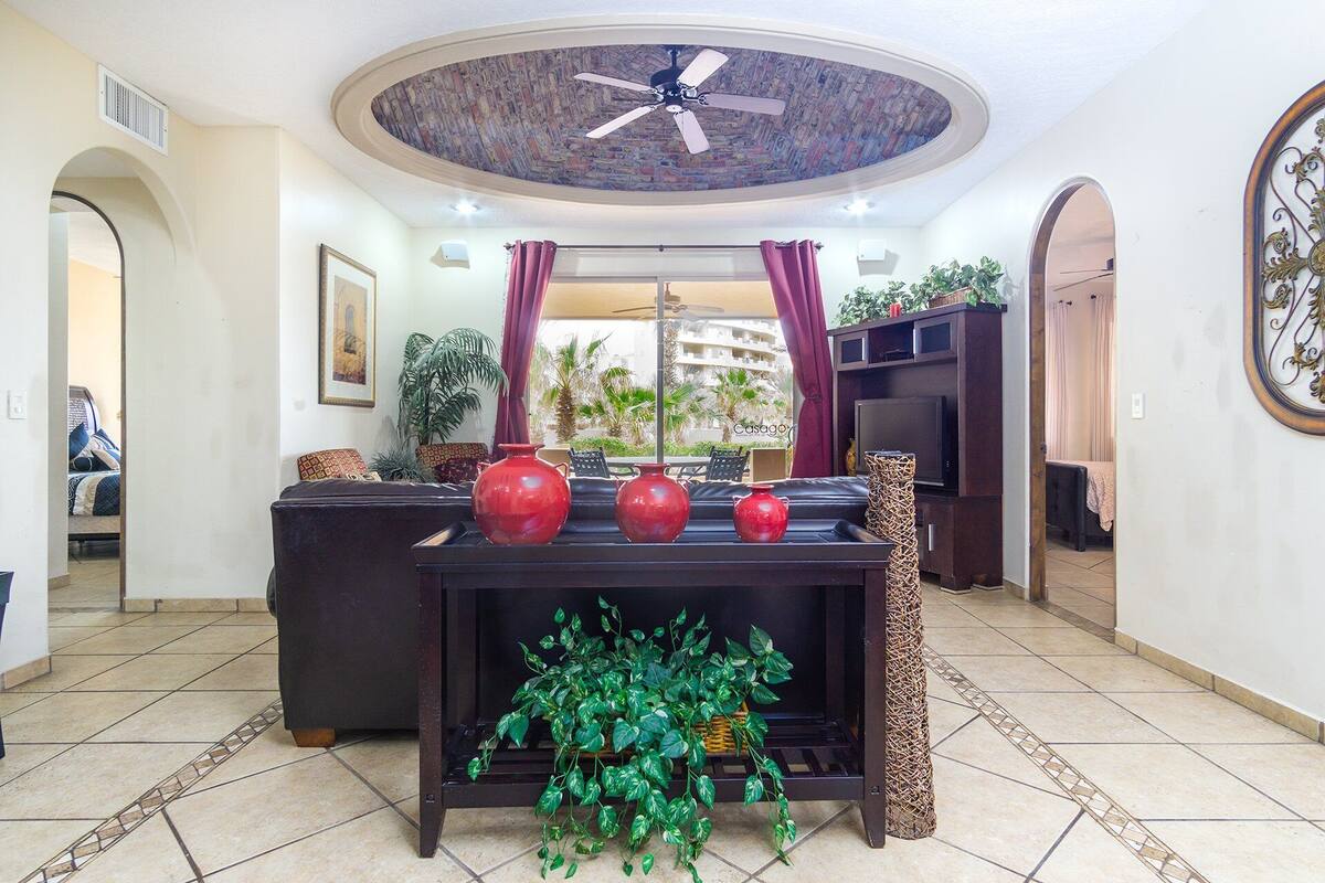 A living room with a ceiling fan, a TV on a wooden stand, and a large window with curtains.