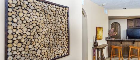 Una sala de estar con una gran obra de arte de madera en la pared, un sofá marrón, una mesita con un jarrón y una cocina con taburetes de madera y un refrigerador.