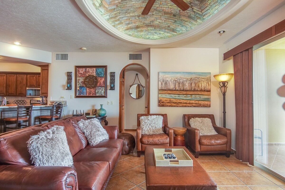 Una sala de estar con un sofá de cuero marrón, dos sillones, una mesa de centro y obras de arte en las paredes. Se ve una cocina al fondo con armarios de madera y un comedor.