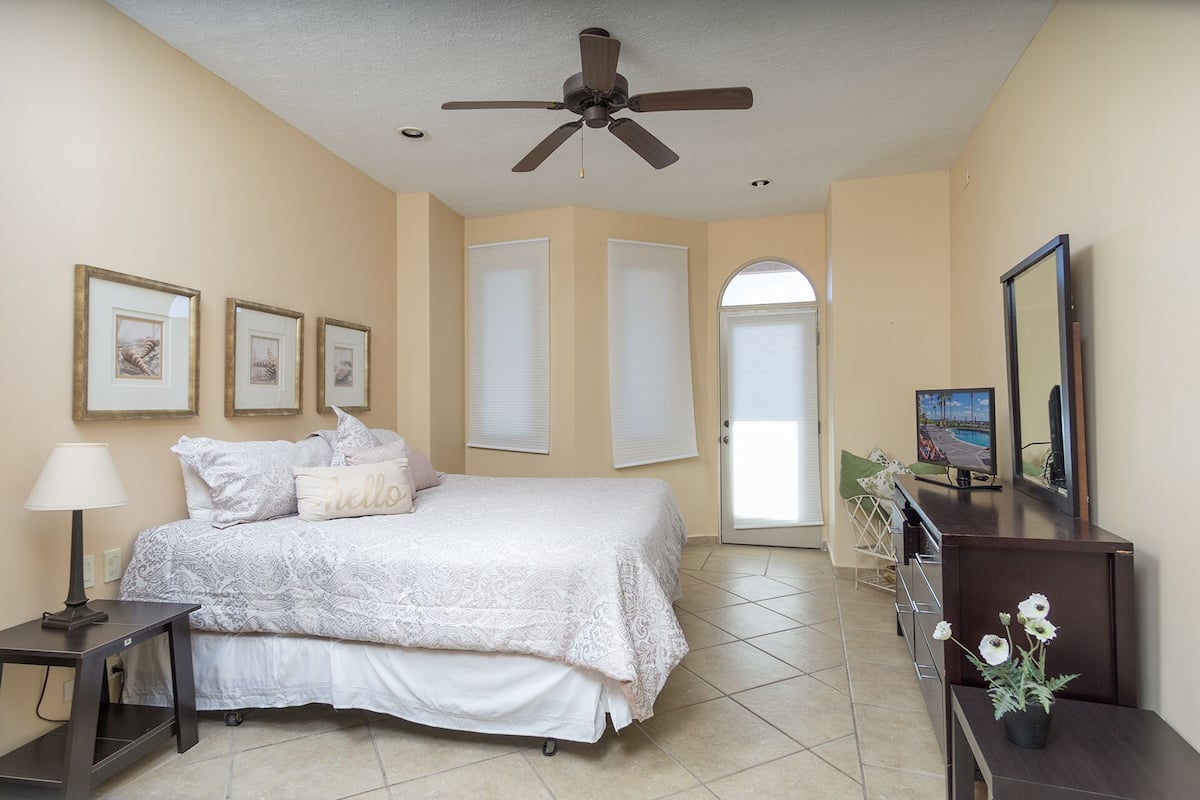 A bedroom with a bed, a ceiling fan, a TV, a mirror, and framed pictures on the wall.