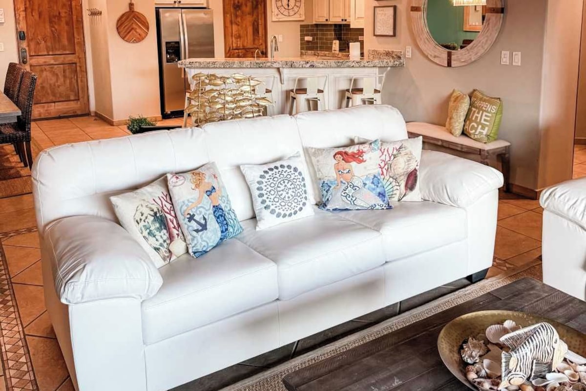 A modern living room with a white sofa, decorative pillows, a coffee table with seashells, a kitchen in the background, and a unique ceiling design.
