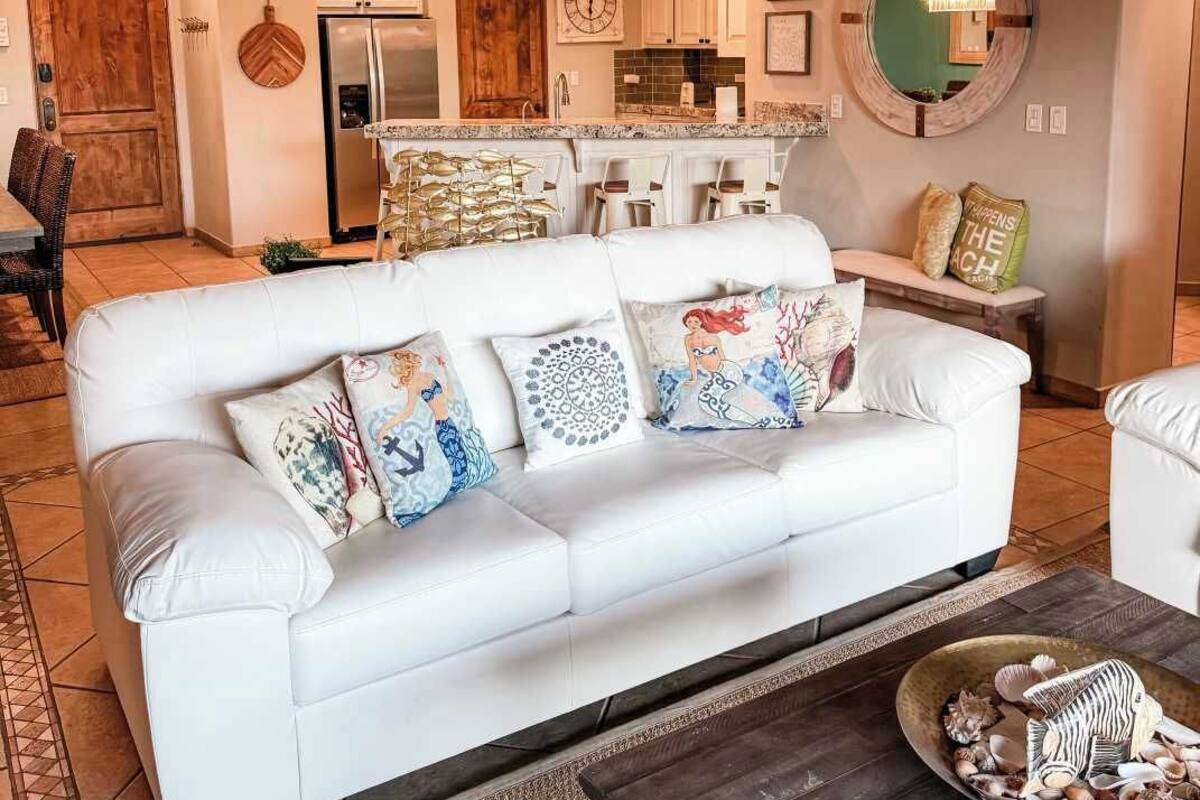 A modern living room with a white sofa, decorative pillows, a coffee table with seashells, a kitchen in the background, and a unique ceiling design.