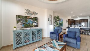 Ein Wohnzimmer mit einem großen Fernseher, zwei blauen Sesseln, einem Couchtisch und einer Anrichte mit Zeitschriften.