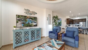 Ein Wohnzimmer mit einem großen Fernseher, zwei blauen Sesseln, einem Couchtisch und einer Anrichte mit Zeitschriften.