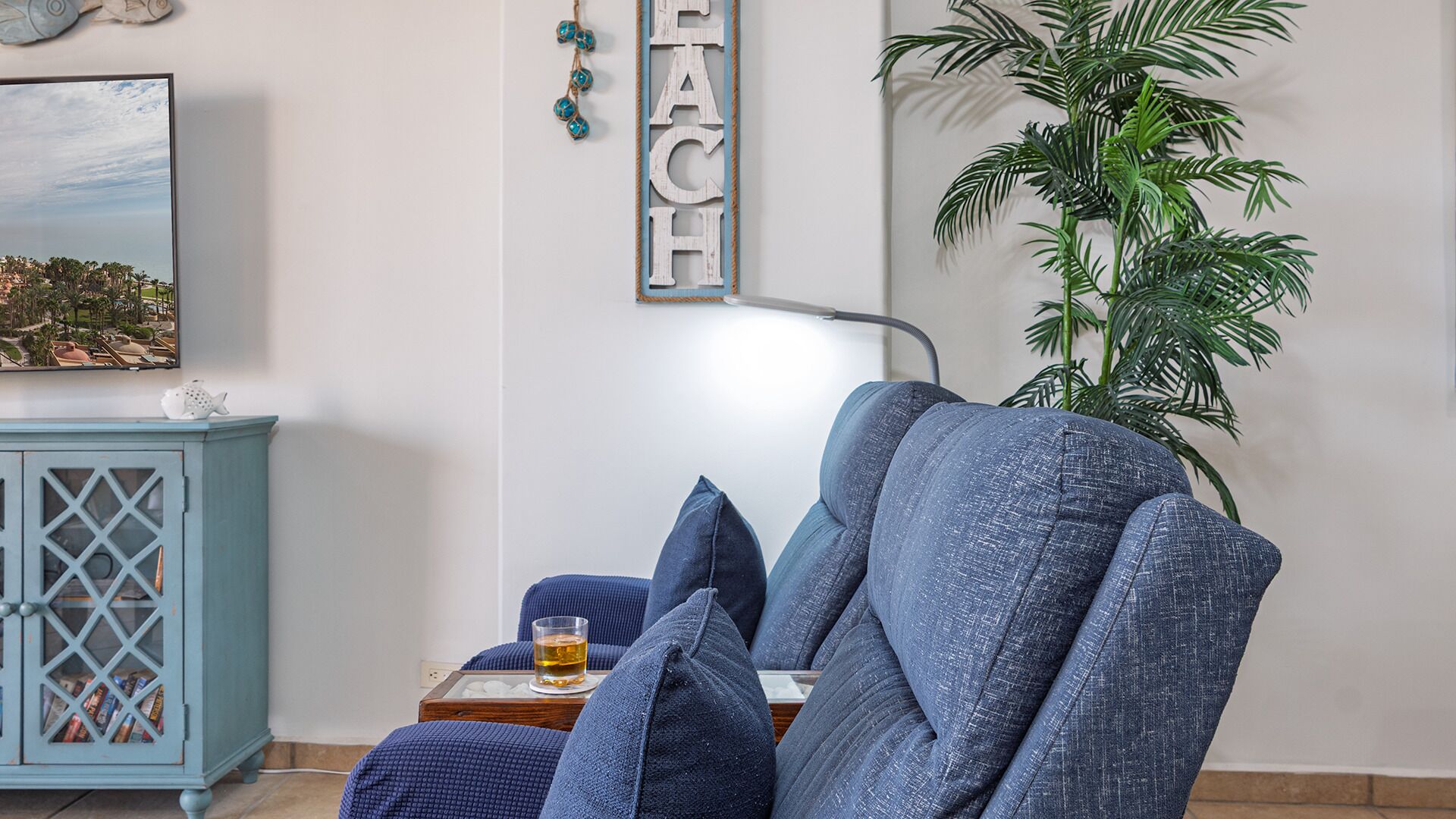 A room with a blue armchair, a small table with a glass of amber liquid, a cabinet with glass doors, a large potted plant, and a wall-mounted sign with the word "BEACH".