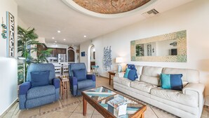 Ein geräumiges Wohnzimmer mit einem weißen Sofa, blauen Sesseln und einem Couchtisch. Im Hintergrund ist eine Küche erkennbar, und an der Wand hängt ein großer Spiegel.