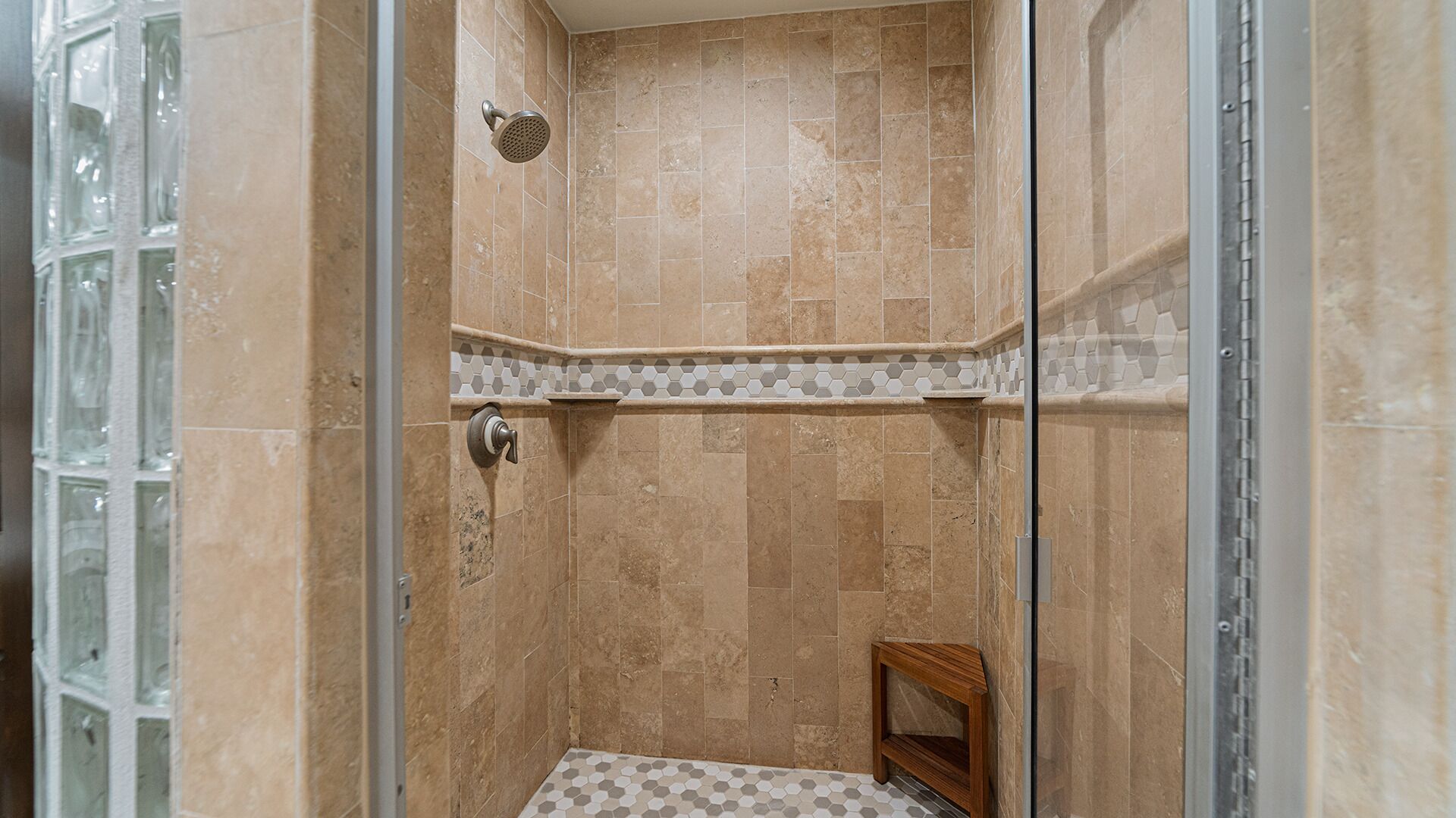 A walk-in shower with beige tiles, a showerhead, and a small wooden stool.