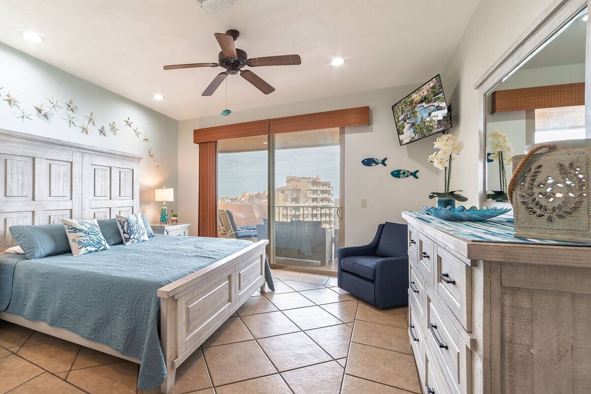A bedroom with a large bed, a ceiling fan, a view of the city, and a balcony with a chair.
