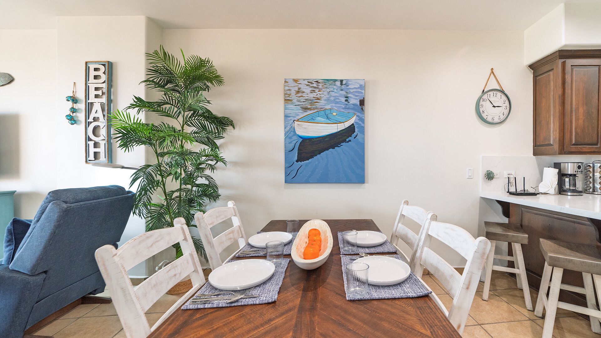 Un coin repas avec une table en bois dressée pour six personnes, entourée de chaises blanches. Une grande plante en pot se trouve à gauche, et un tableau représentant un bateau sur l’eau est accroché au mur.