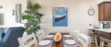 Un coin repas avec une table en bois dressée pour six personnes, entourée de chaises blanches. Une grande plante en pot se trouve à gauche, et un tableau représentant un bateau sur l’eau est accroché au mur.