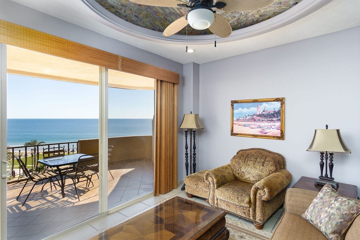 A living room with a sofa, a coffee table, and a painting on the wall. There is a balcony with a view of the ocean.