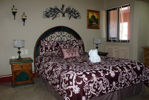 A bedroom with a large bed, a nightstand, a wall-mounted lamp, a decorative wall piece, a window with bars, and a painting. - Marina Pinacate (Puerto Peñasco)