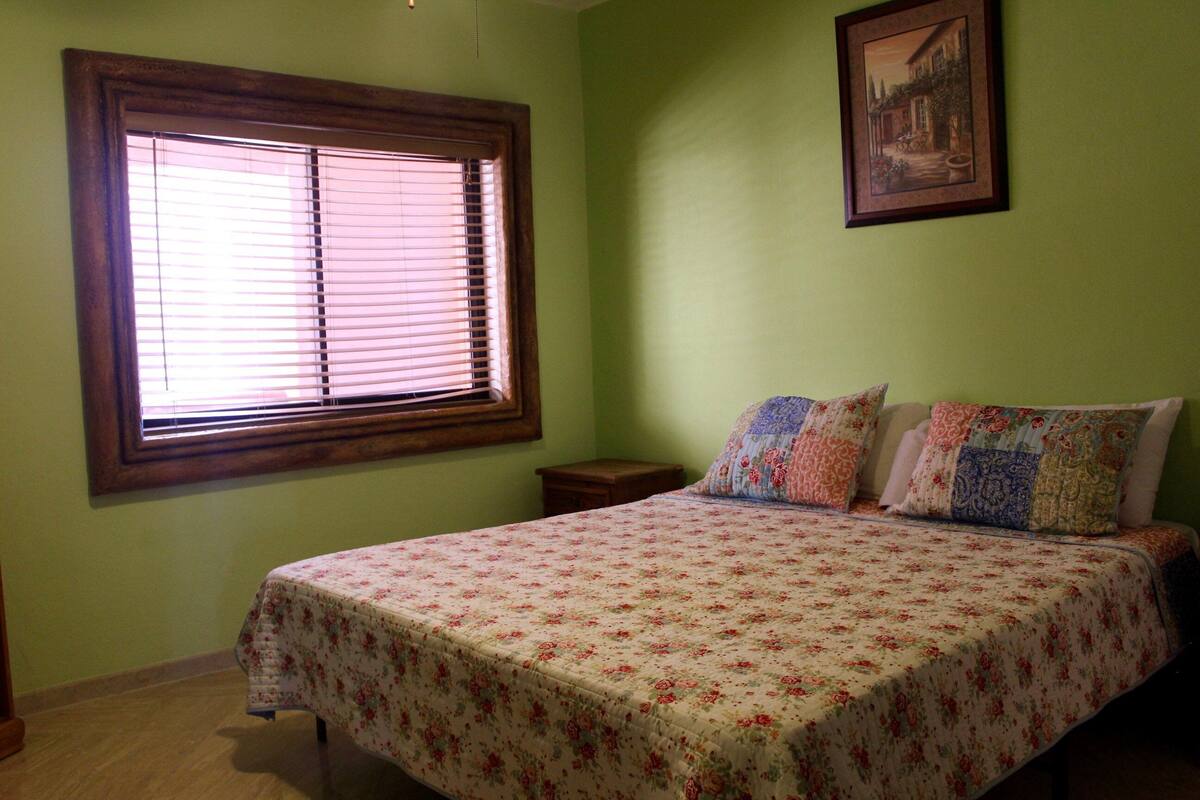 A bedroom with a bed, a nightstand, a window with blinds, and a framed picture on the wall.