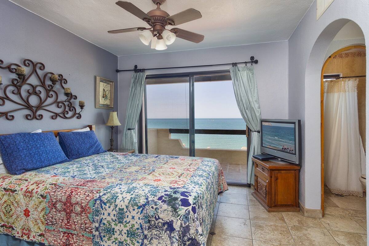 A bedroom with a bed, a ceiling fan, a television, and a view of the sea.