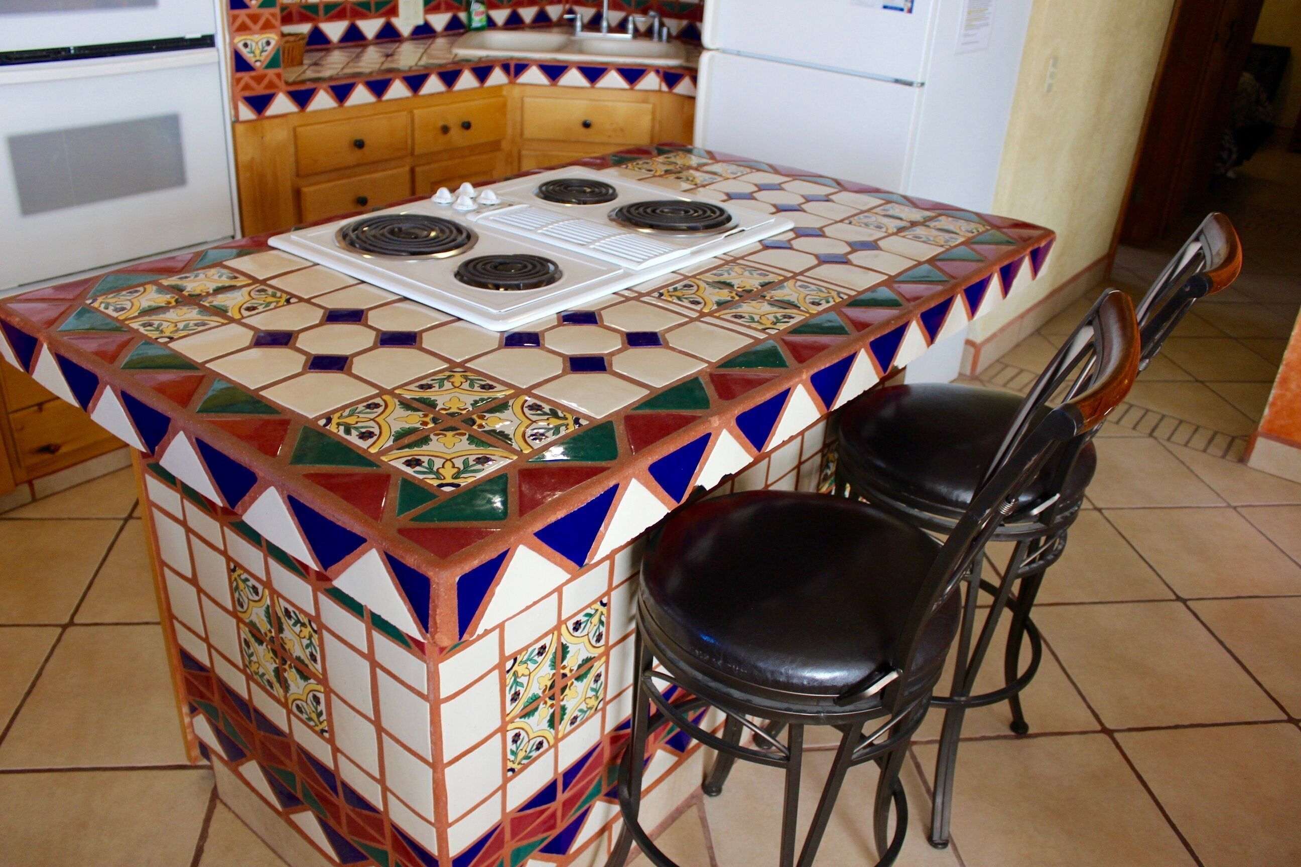 A kitchen with a tiled island, a stove, and two black chairs.