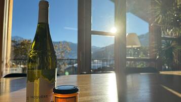 Une bouteille de vin et un pot de miel sont posés sur une table en bois, d’où l’on peut admirer un paysage de montagnes et un plan d’eau à travers de grandes fenêtres.