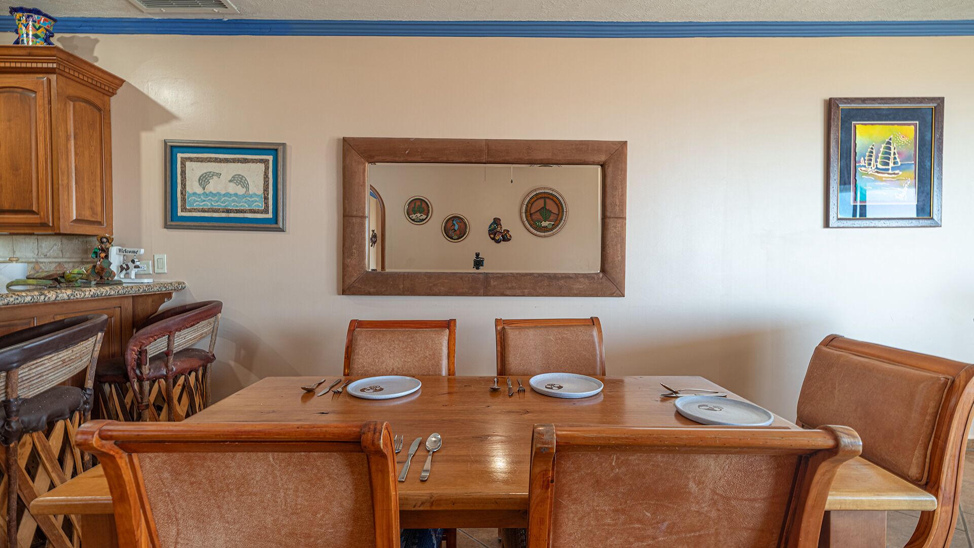 A dining area with a wooden table and chairs, framed artwork on the wall, and a kitchen area with wooden cabinets and a welcome sign.