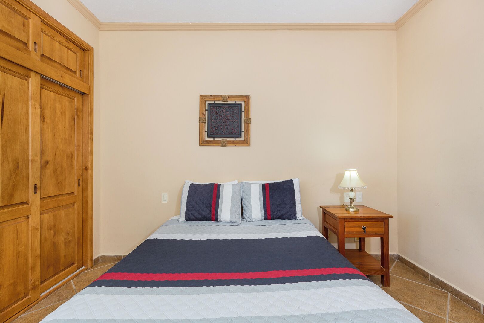Um quarto com uma cama, uma mesa de cabeceira com abajur, um guarda-roupa de madeira e um quadro na parede.