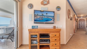 Una sala de estar con un televisor mostrando una escena de playa, un mueble de madera para entretenimiento con dispositivos electrónicos y un letrero que dice “THE OCEAN FIXES EVERYTHING”.