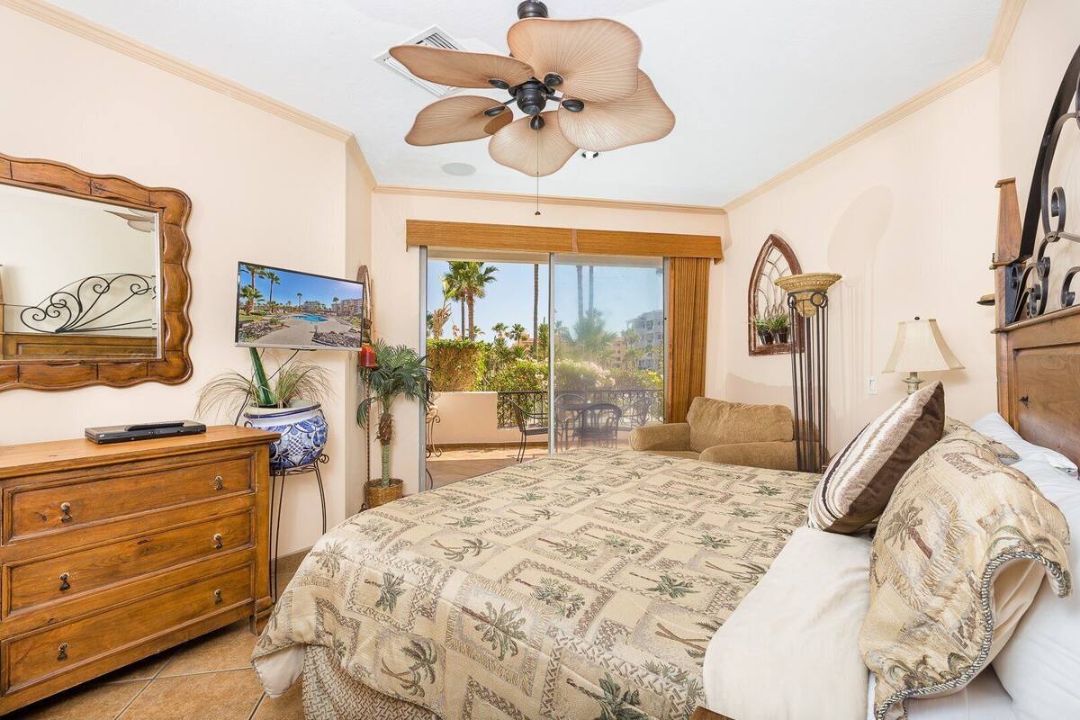 Ein Schlafzimmer mit einem großen Bett, einem Deckenventilator, einem Fernseher, einem Schminktisch und einem Balkon mit Blick auf Palmen.