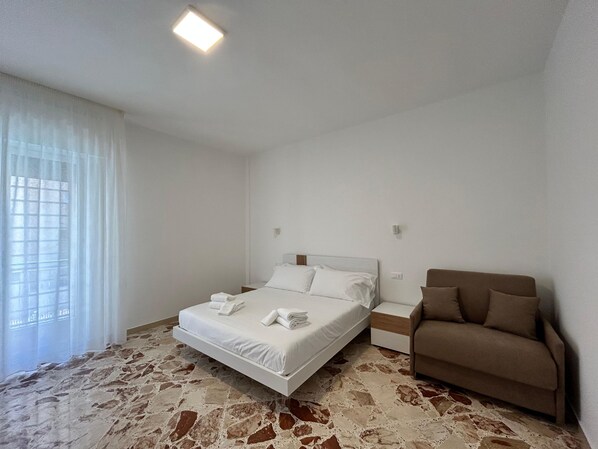 A bedroom with a bed, a nightstand, a brown sofa, and a window with sheer curtains.